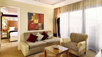 A living room with a sofa, armchair, and a coffee table. There is a painting on the wall and a large window with curtains.