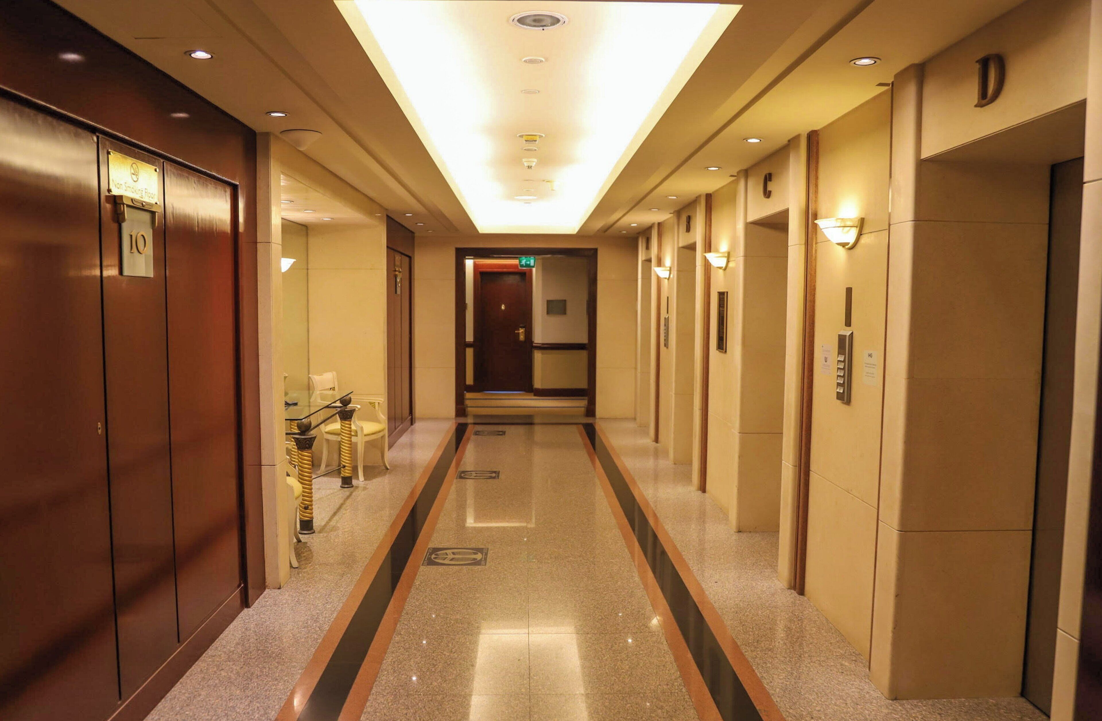 A hotel corridor with doors on both sides, a central walkway, and a glass partition with chairs.
