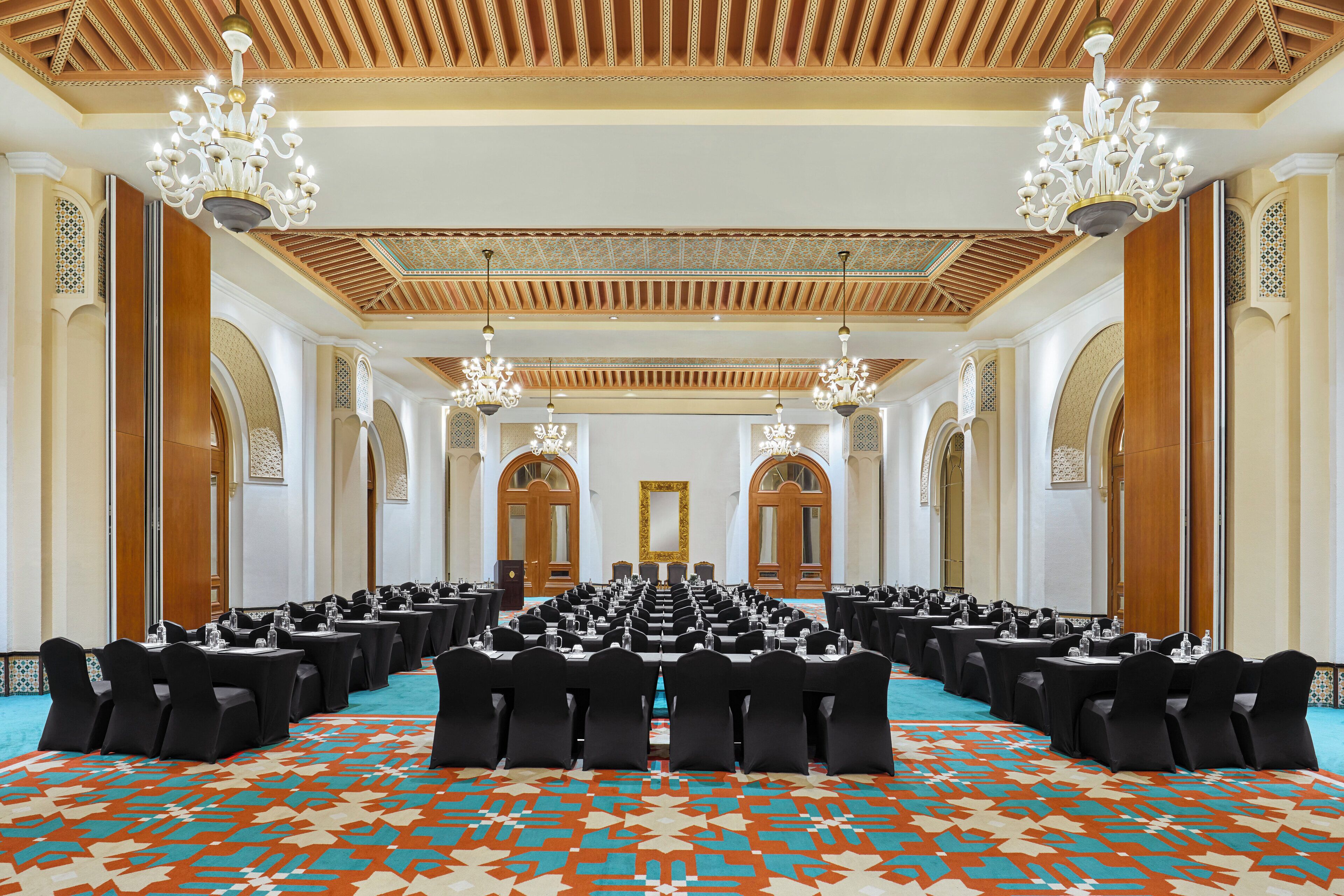 A conference room with rows of tables and chairs, chandeliers, and ornate ceiling design.