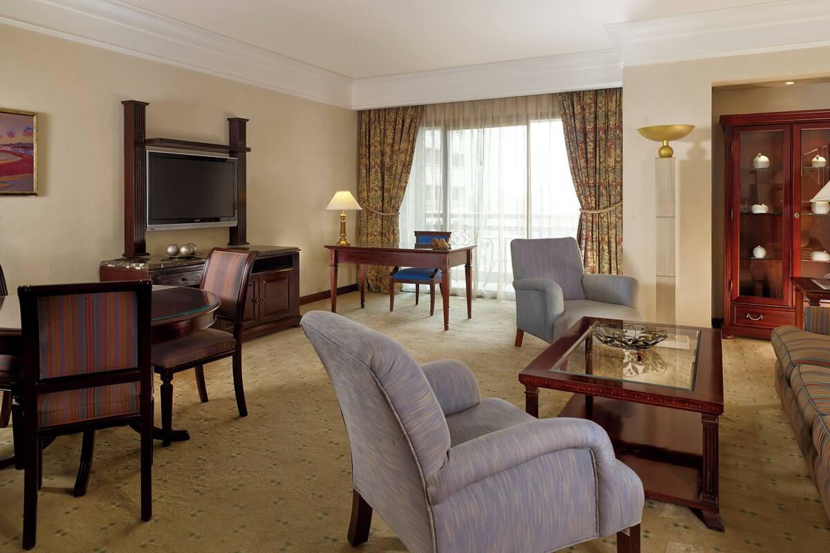 A hotel room with a flat-screen TV, a dining table, two armchairs, a sofa, a cabinet with glass doors, and a painting on the wall.