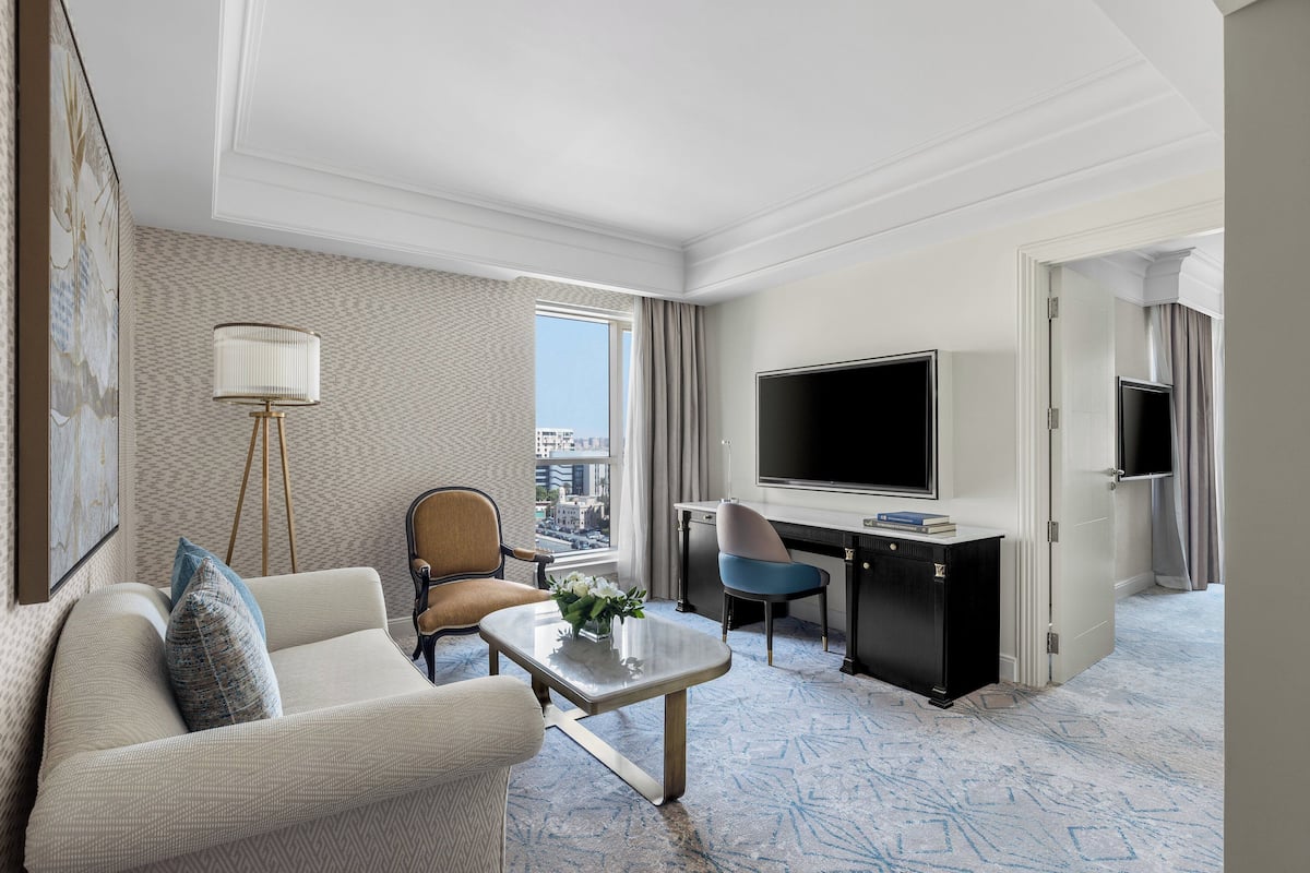 A hotel room with a sofa, armchair, desk, TV, and a view of the city.