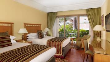 A hotel room with two beds, a sitting area with a table and chairs, a TV, and a balcony with plants.