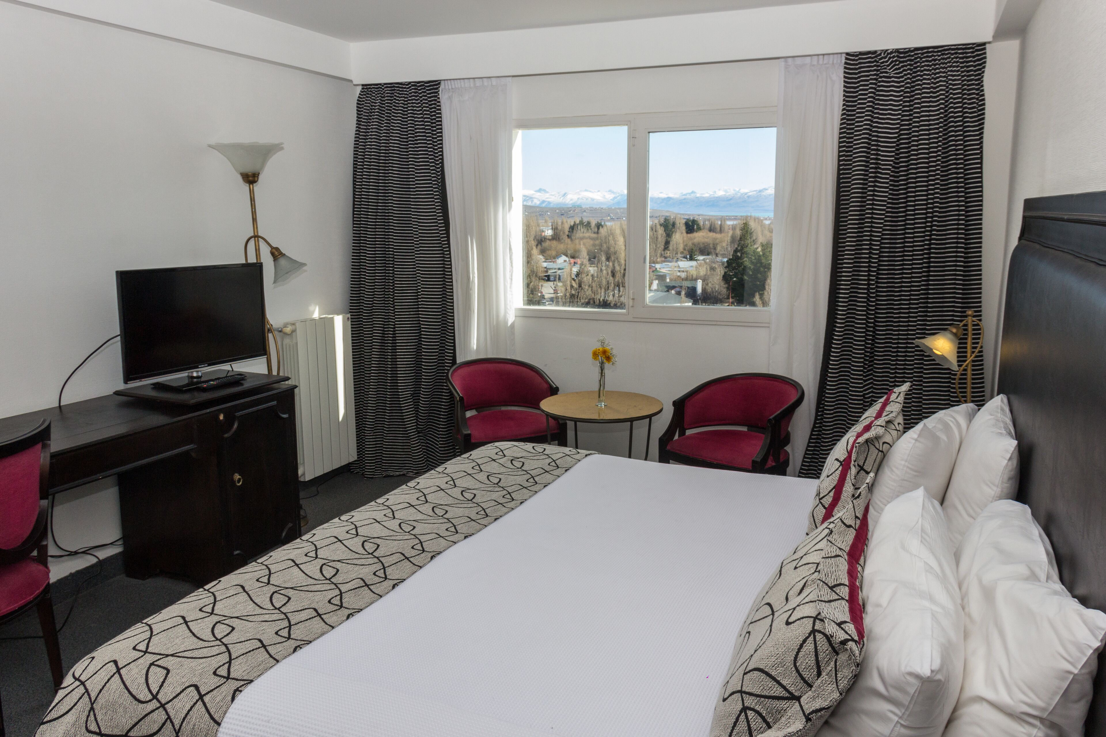 A hotel room with a bed, a desk, a TV, two chairs, a small table, and a window with a view of mountains.