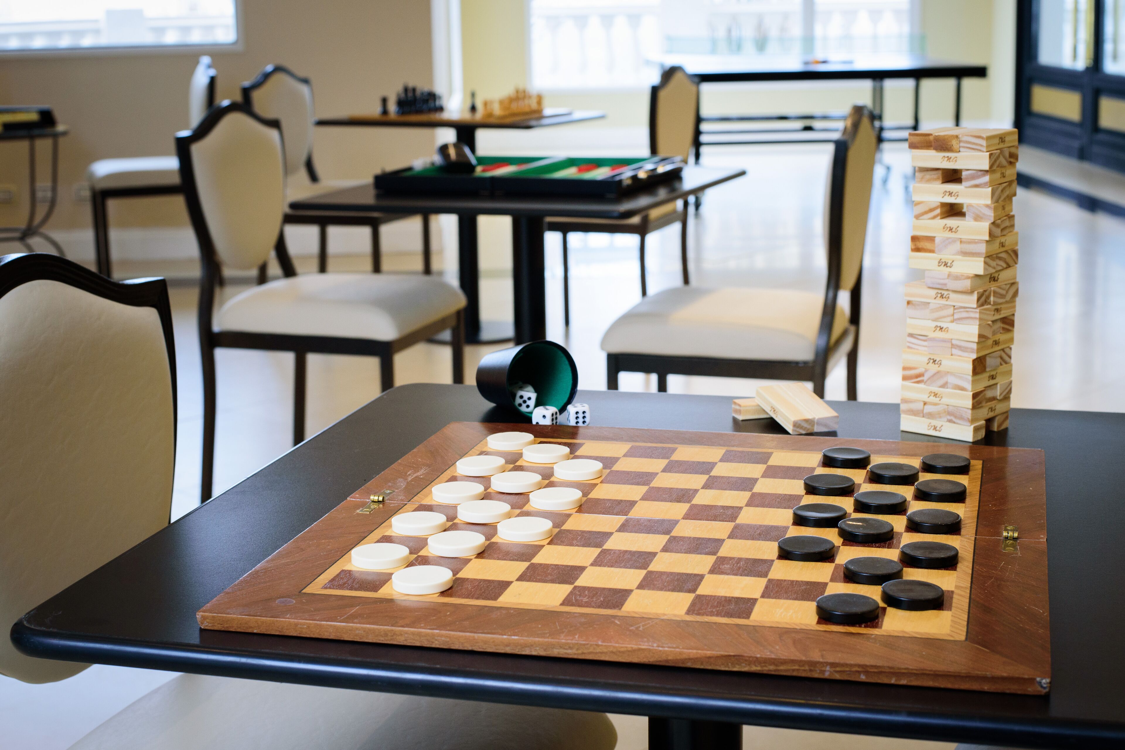 A checkered board game with dice and wooden blocks on a table in a room with chairs and tables.
