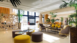 A spacious lobby with a modern design, featuring a seating area with ottomans and a sofa, a chandelier, and large windows allowing natural light.