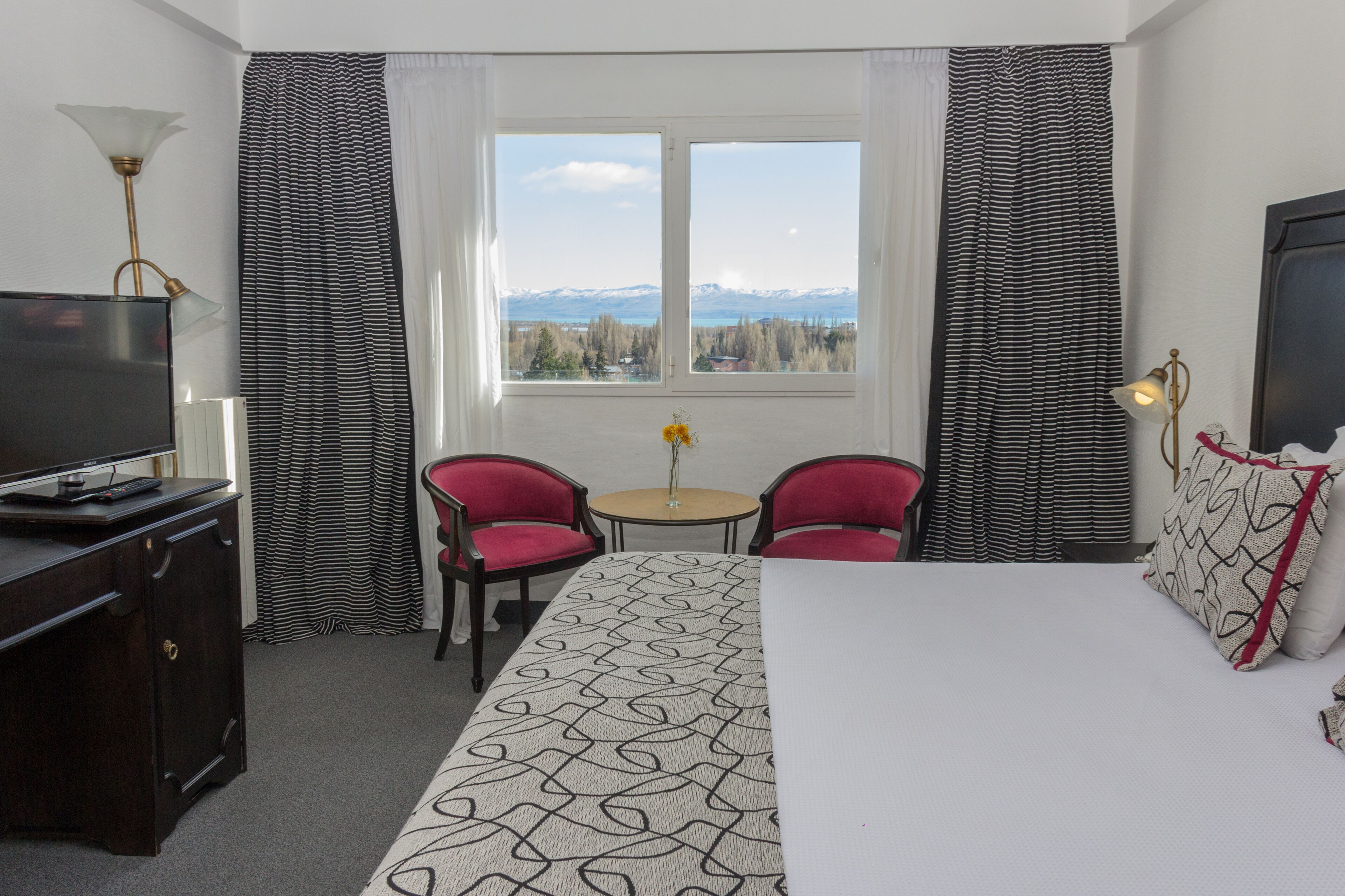 A hotel room with a bed, a desk with a TV, two red chairs, a small table with a vase, and a window with a mountain view.