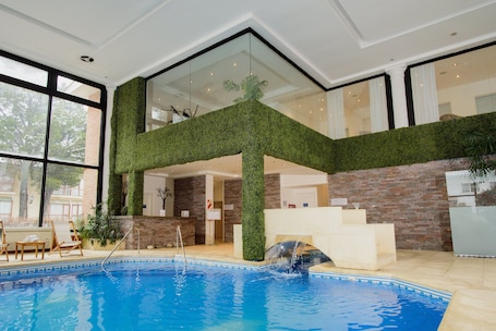 An indoor swimming pool with a glass ceiling, surrounded by brick walls and a green artificial grass wall.