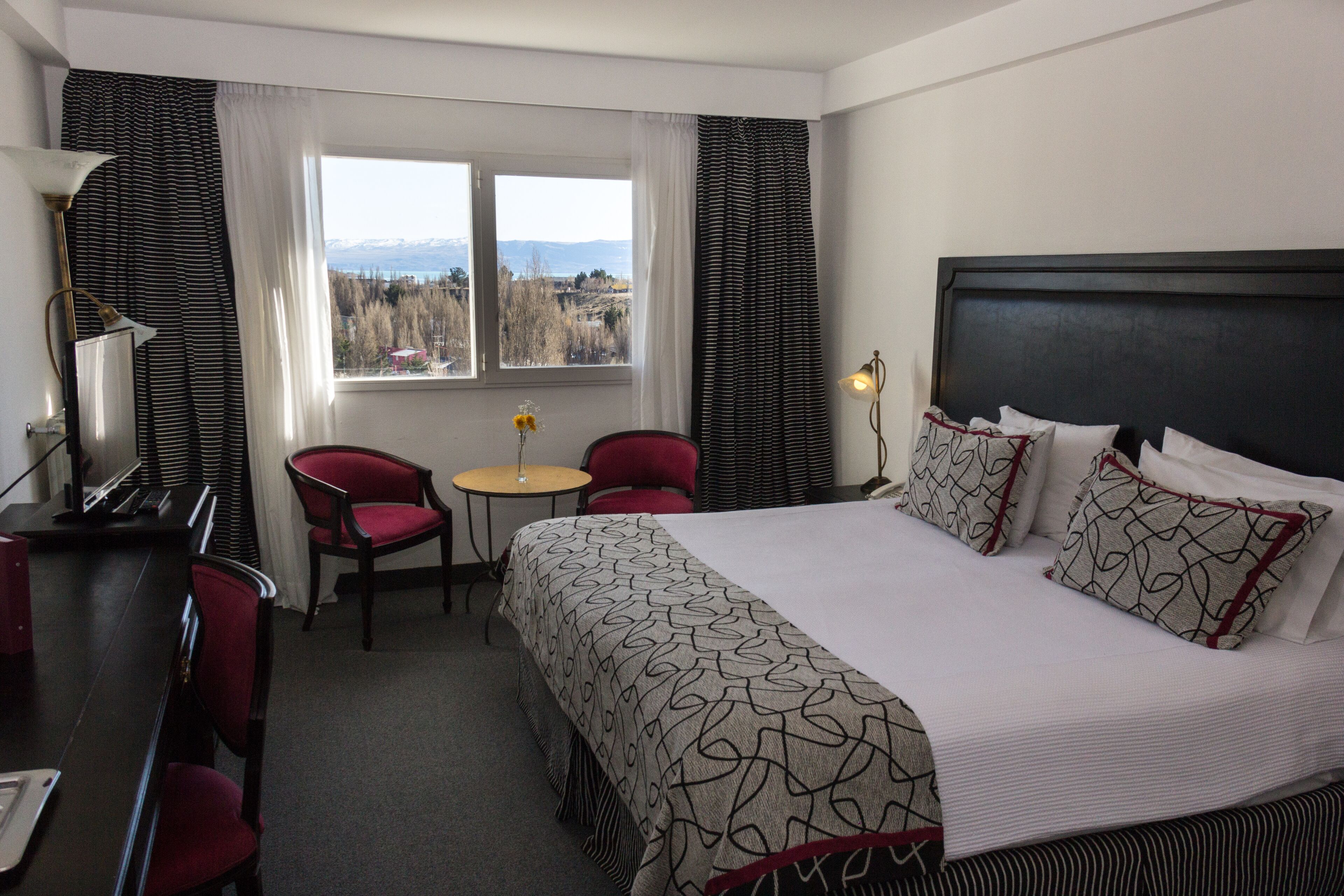 Habitación de hotel con una cama grande, un escritorio, dos sillas, una mesita y una ventana con vistas a montañas.