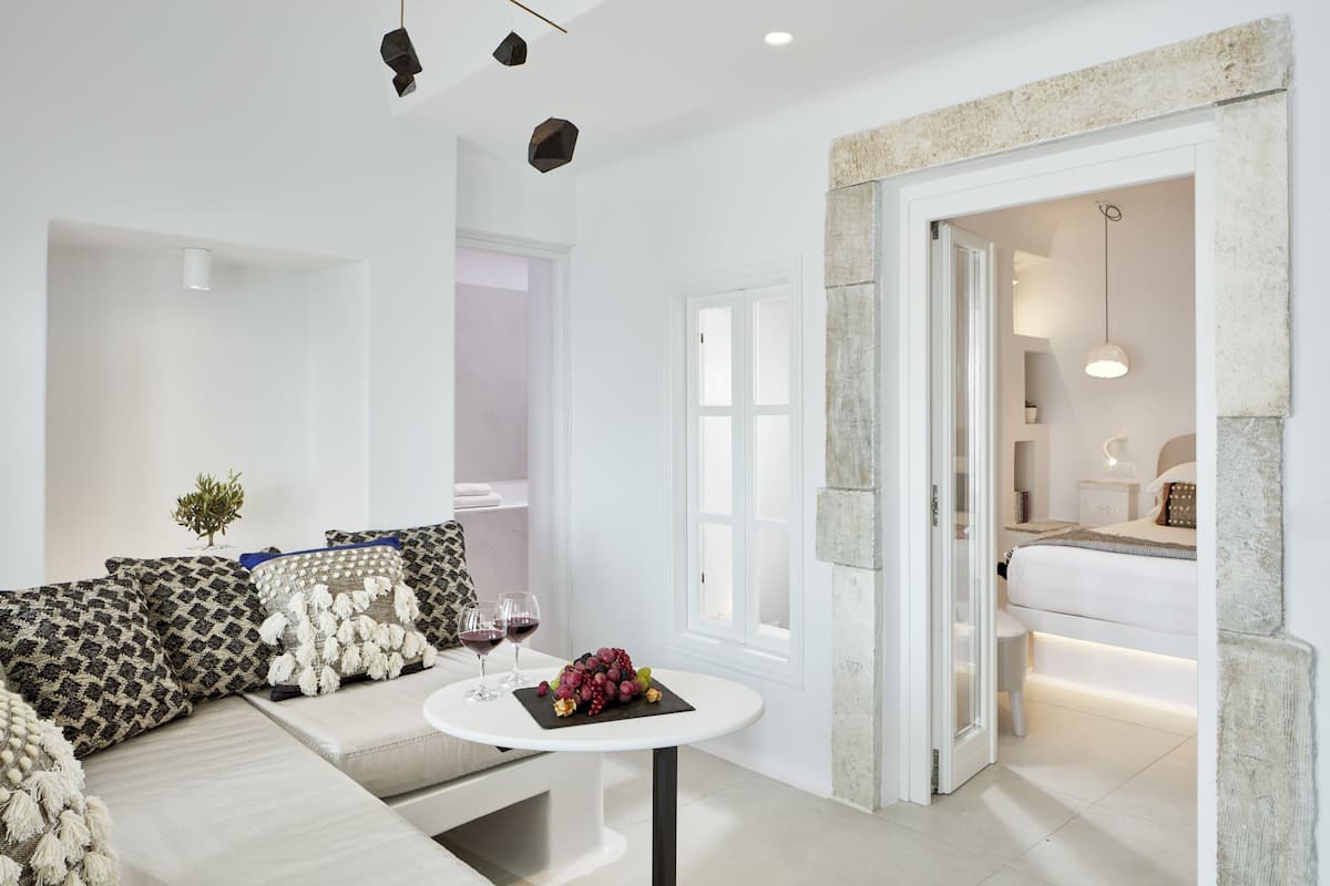 A modern living room with a sofa, a round table with a fruit bowl, and a doorway leading to a bedroom.