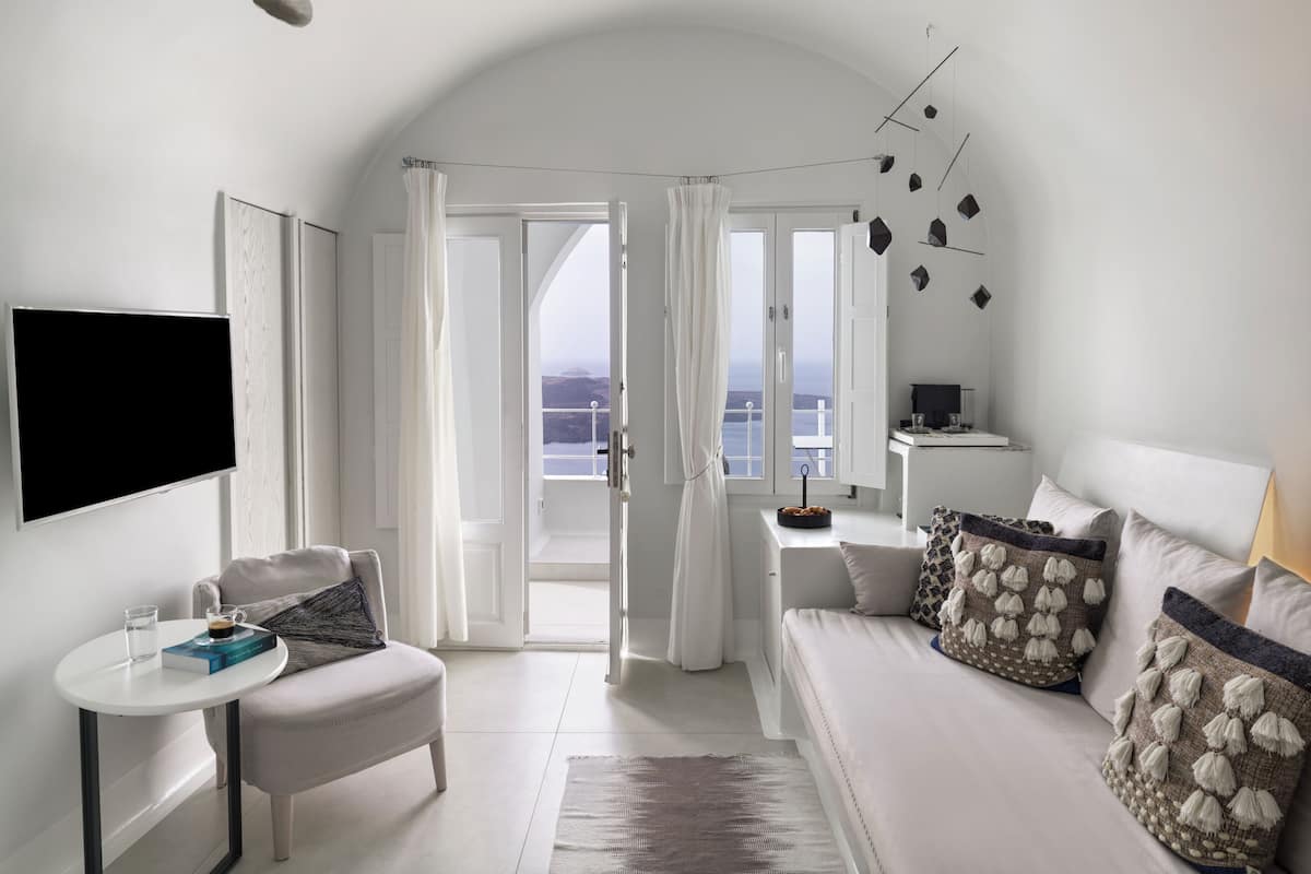 A modern hotel room with a balcony, a flat-screen TV, a small table with a book and a glass of water, and a sofa with decorative pillows.