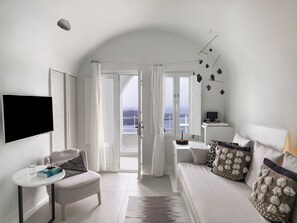 Una habitación de hotel moderna con balcón, televisión de pantalla plana, una mesita con un libro y un vaso de agua, y un sofá con cojines decorativos.