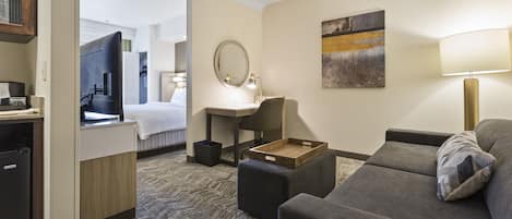 A hotel room with a grey sofa, a wooden side table, a desk with a lamp, a television, and a bed with a white headboard.