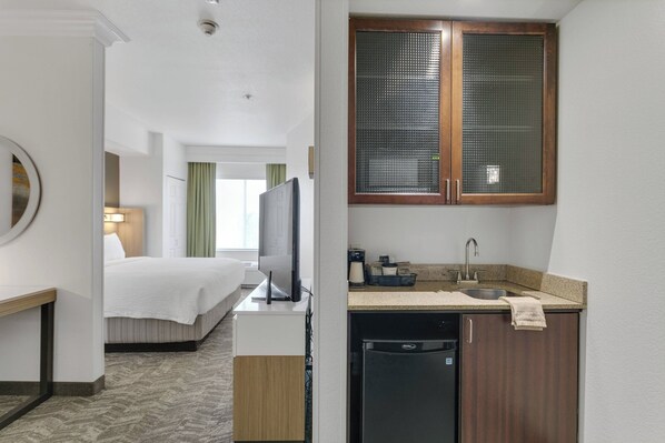 A hotel room with a bed, a desk, a television, a kitchen area with a sink and a dishwasher, and a large window with blinds. - SpringHill Suites by Marriott Sacramento Natomas (Sacramento)