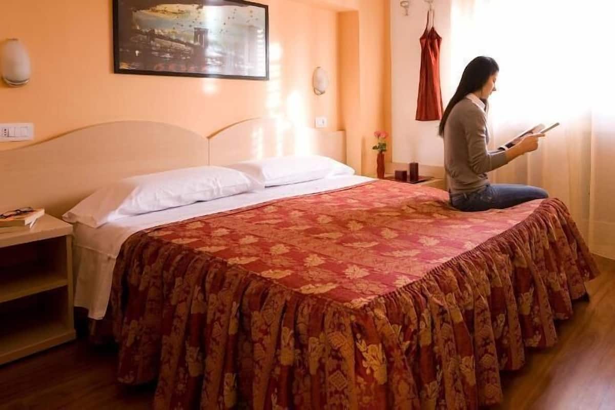 A bed with a red floral bedspread, a nightstand with a book, a wall-mounted lamp, and a painting above the bed.