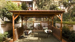 A covered patio area with tables and chairs, surrounded by greenery and a fence. - Hotel Sabatino (Milan)