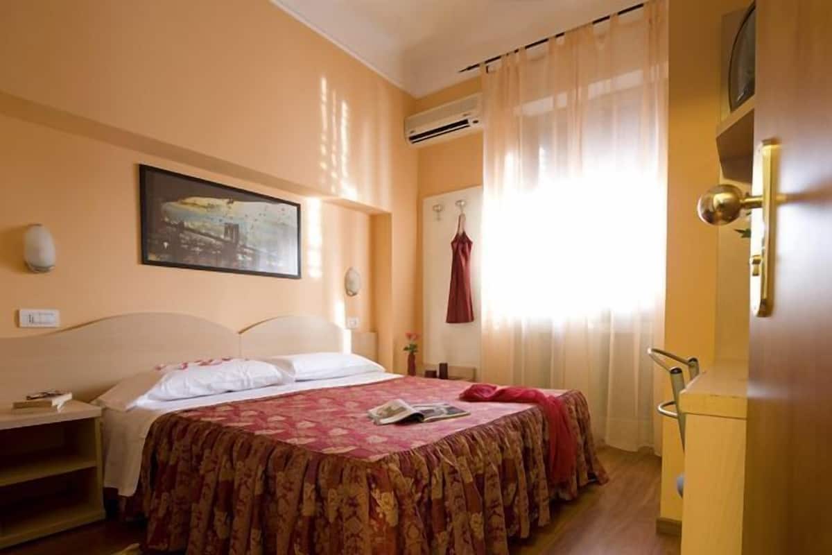 A hotel room with a bed, a nightstand, a window with curtains, an air conditioning unit, and a painting on the wall.