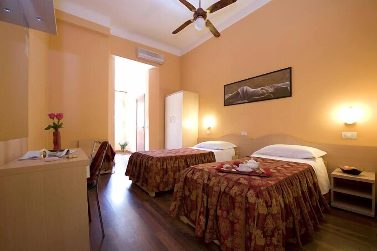 A hotel room with two beds, a desk, a ceiling fan, and a painting on the wall.