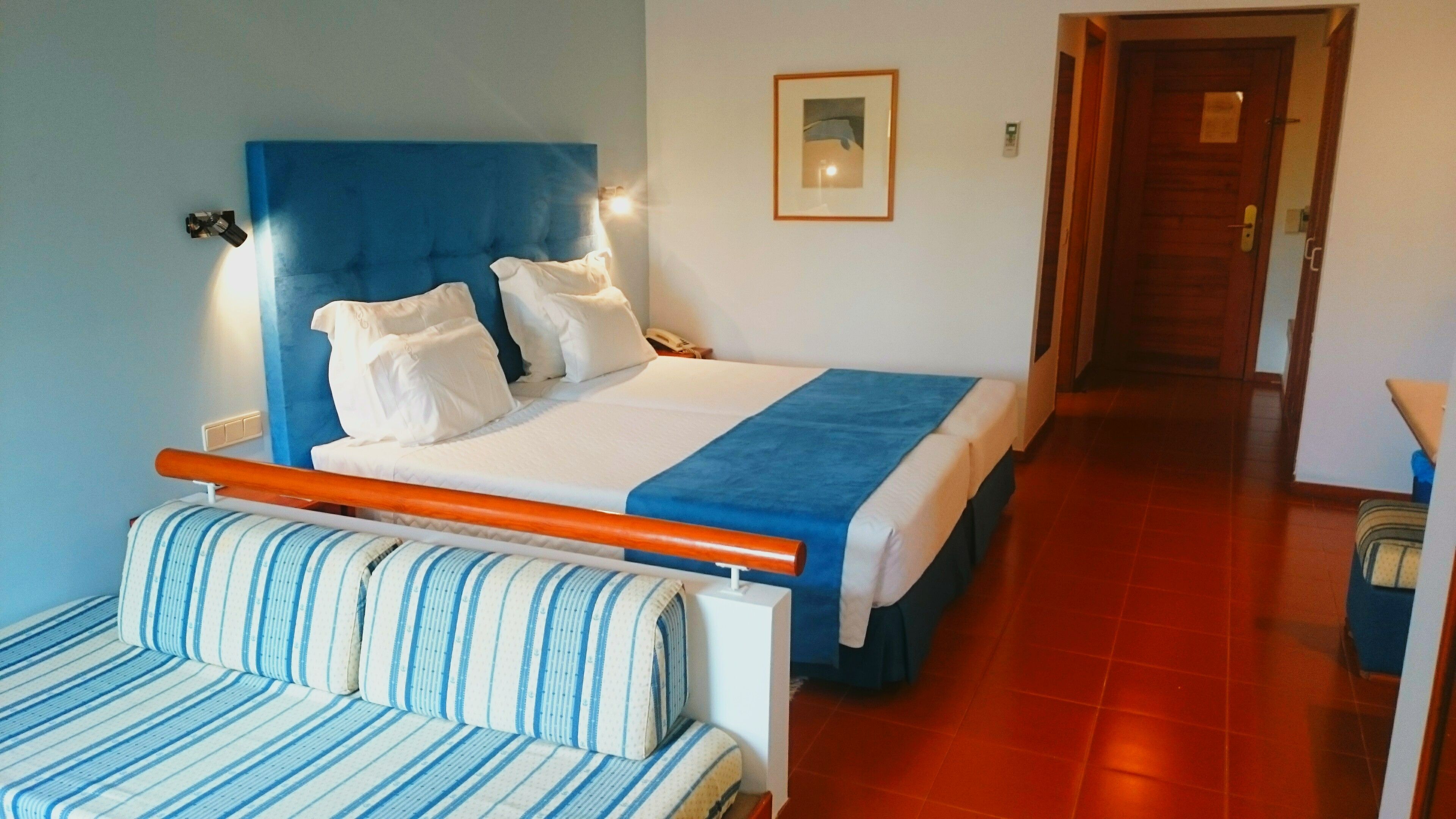 Ein Bett mit blauer Kopfteil, weißer Bettwäsche und einer blauen Tagesdecke. Im Vordergrund steht ein gestreiftes Sofa und im Hintergrund eine Holztür.
