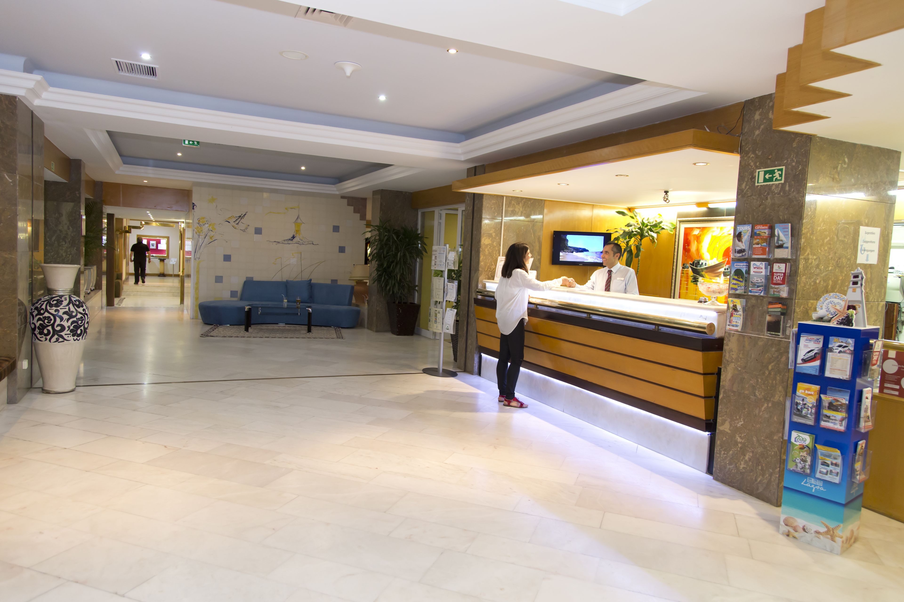 A hotel reception area with a reception desk, a TV, a blue seating area, and a plant.