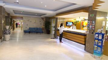 A hotel reception area with a reception desk, a TV, a blue seating area, and a plant.