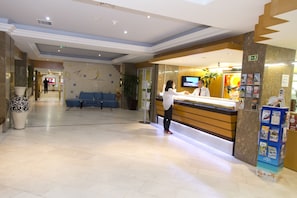 A hotel reception area with a reception desk, a TV, a blue seating area, and a plant.