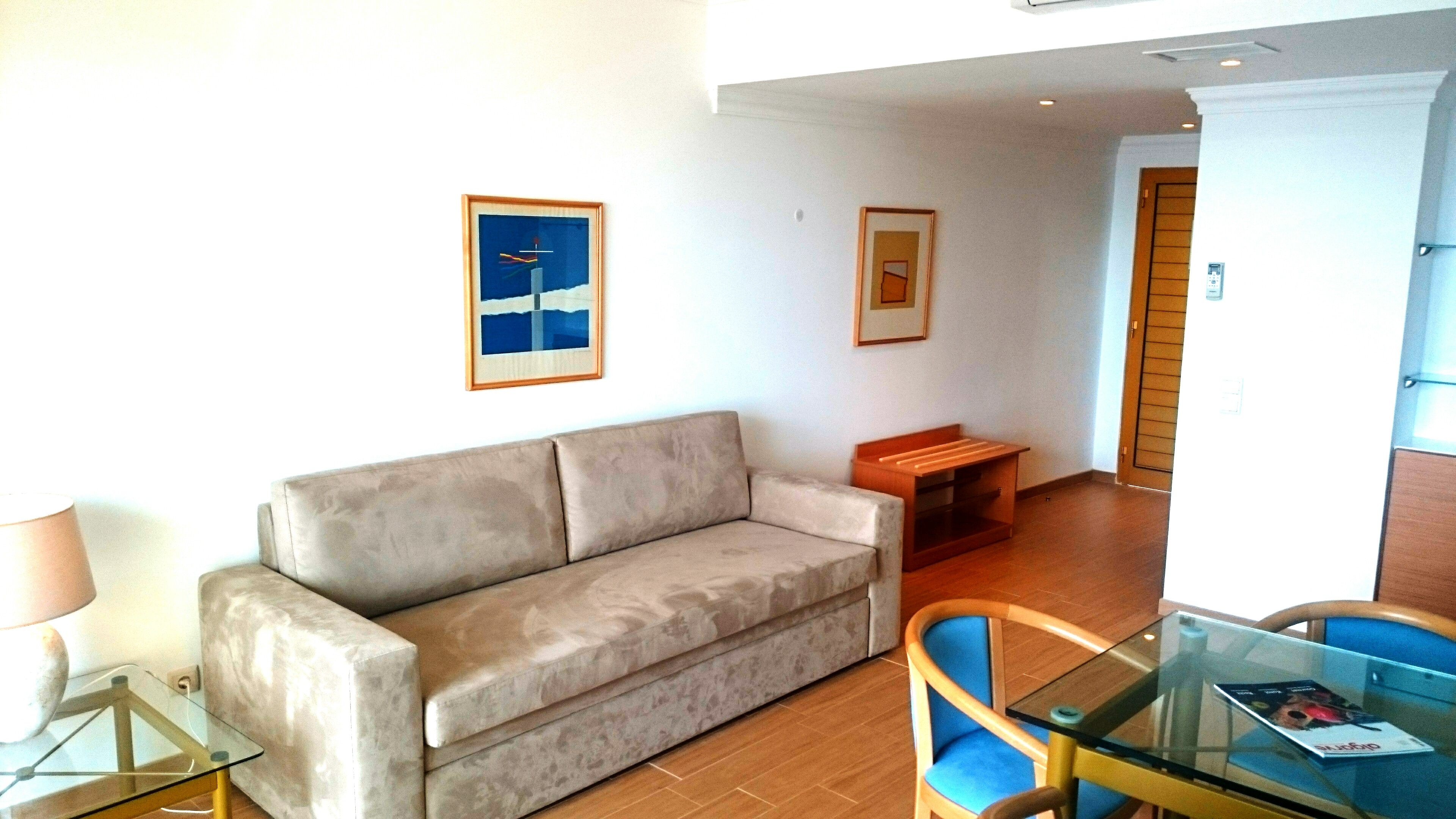 A living room with a sofa, a glass coffee table, and a chair. There are framed pictures on the wall.