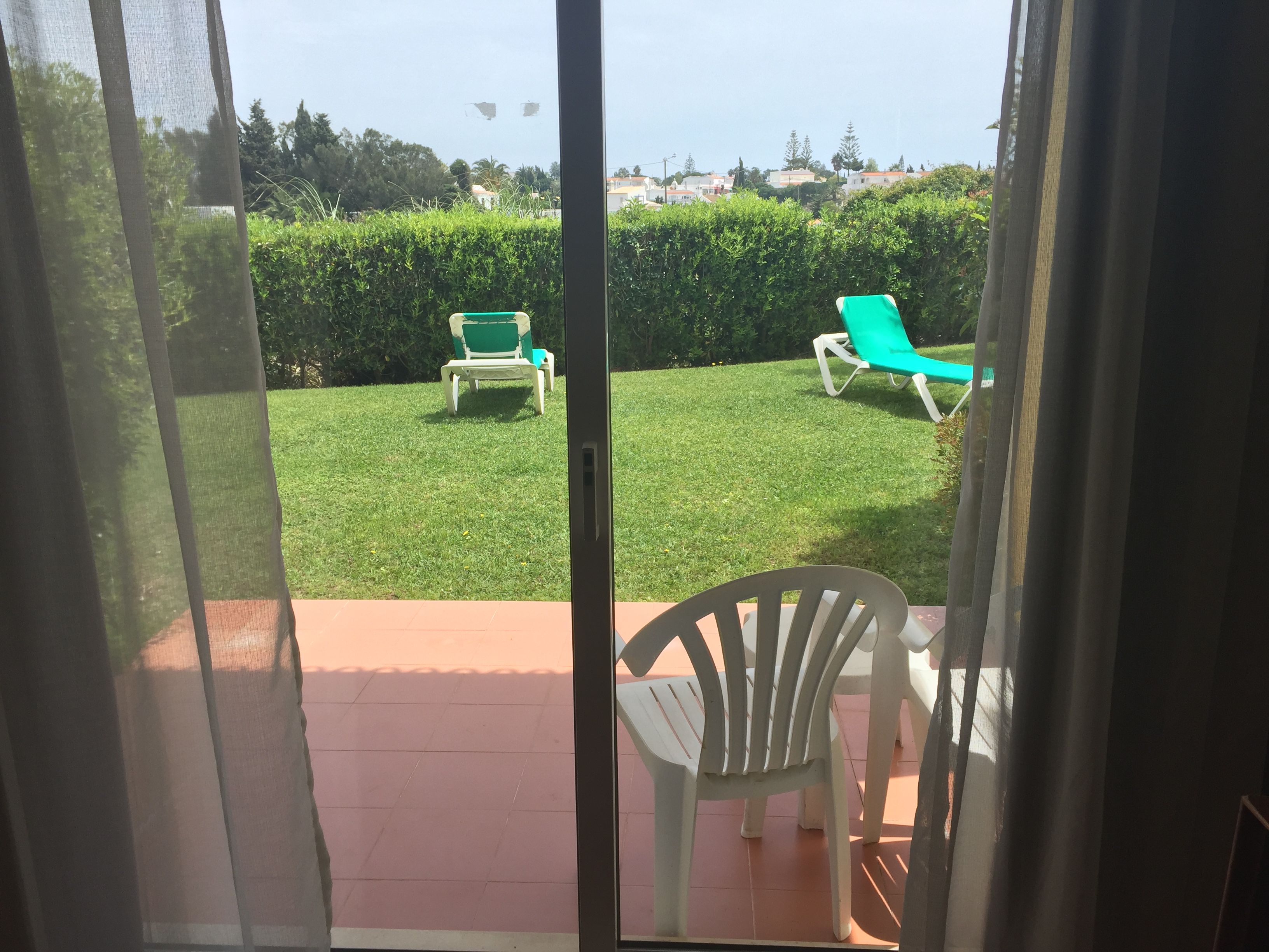 A view through a glass door of a patio area with a lawn, two green lounge chairs, and a white chair.