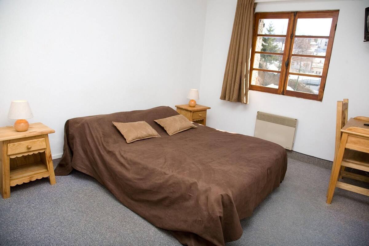 A bedroom with a bed, two bedside tables with lamps, a radiator, a window with curtains, and a chair.