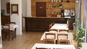 A dining area with wooden tables and chairs, a kitchen area with shelves of bottles and a coffee machine, and a wooden door. - Adonis Le Chastellan (Guillaumes)