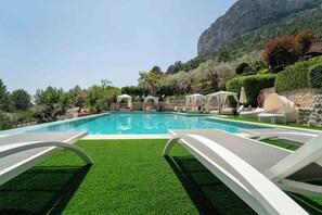 A swimming pool with lounge chairs and umbrellas, surrounded by greenery and a mountain backdrop.