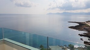 A panoramic view from a balcony overlooking a calm sea with distant mountains, a rocky shoreline, and a clear sky.