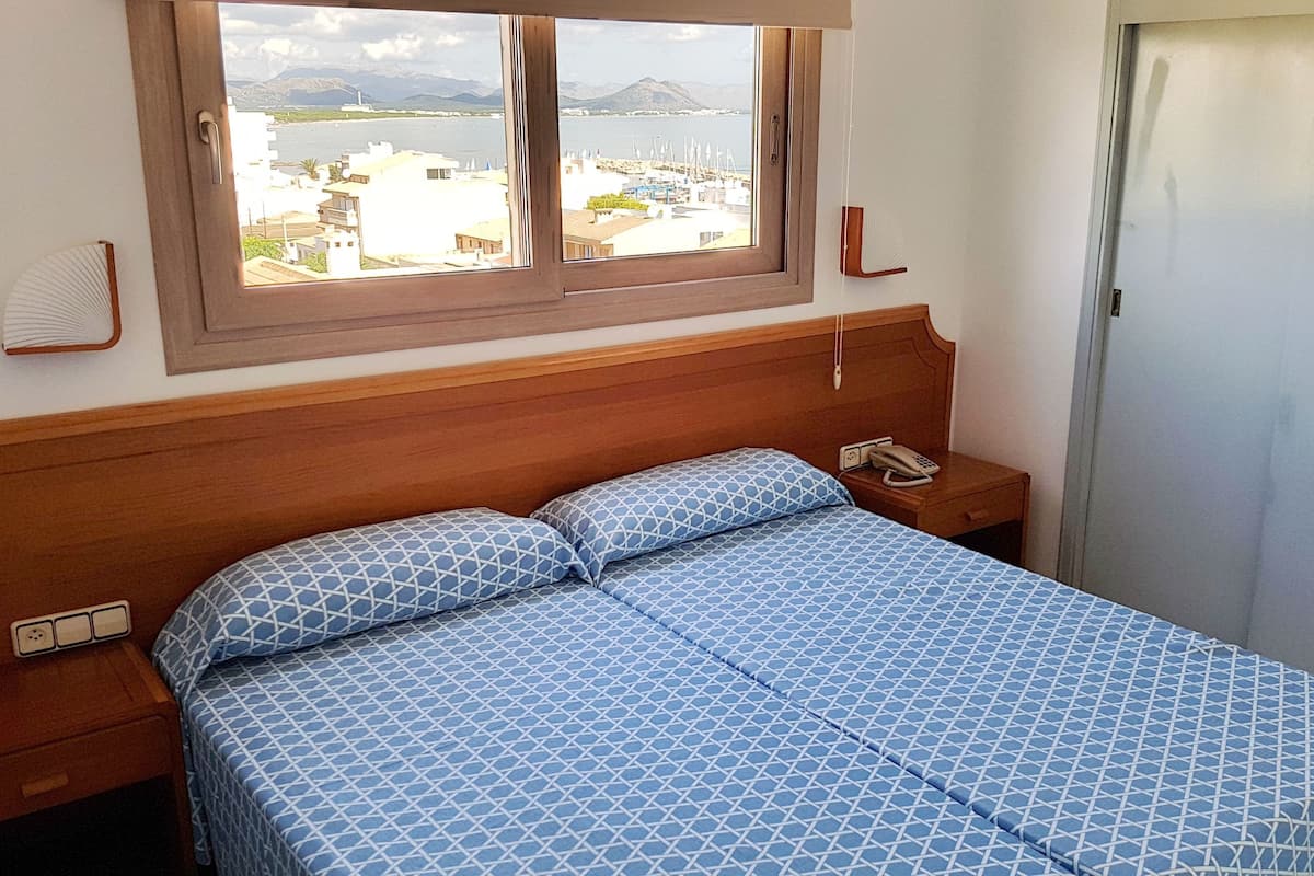 Een hotelkamer met een bed, een nachtkastje met een telefoon, een raam met uitzicht op water en gebouwen, en een deur.