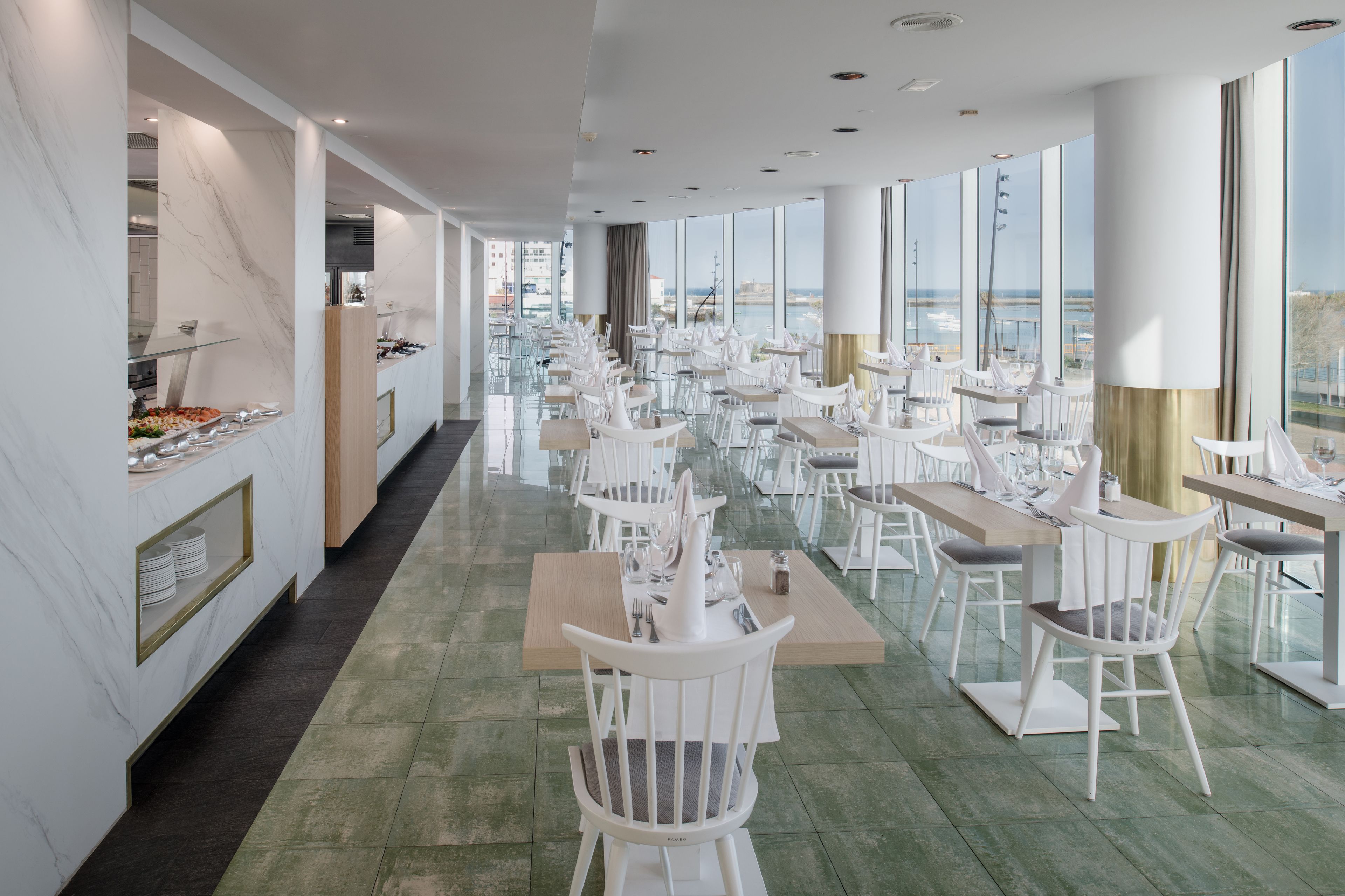 A modern dining area with white chairs and tables, a buffet on the left, and large windows offering a view of the sea.