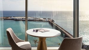 A room with a large window overlooking a pool and the sea. There is a round table with a magazine and sunglasses on it. - Arrecife Gran Hotel & Spa (Arrecife)