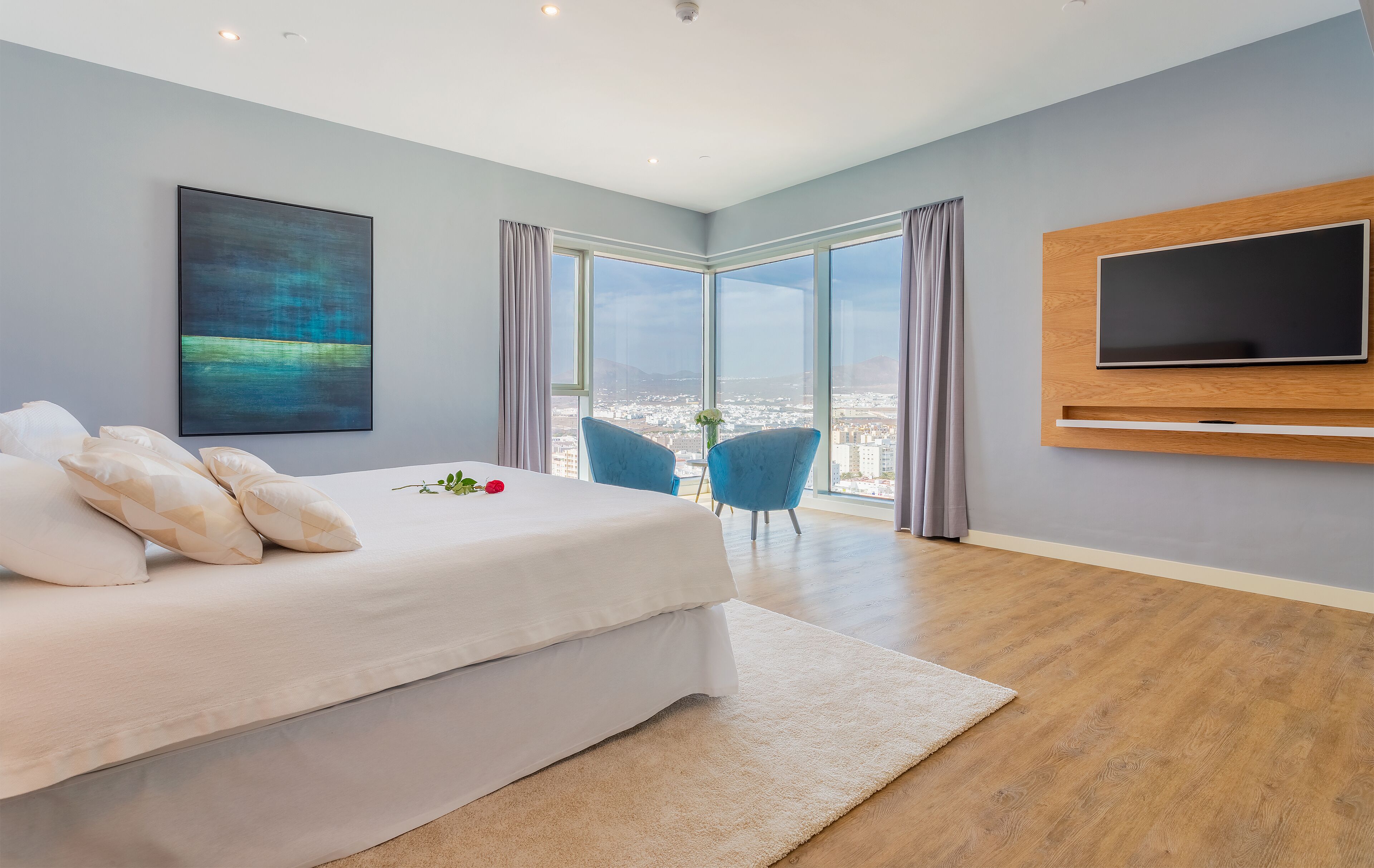 A modern bedroom with a large bed, a mounted TV, and a city view through large windows.