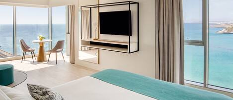 A modern hotel room with a large bed, a TV mounted on the wall, a balcony with a view of the sea, and a small dining area with chairs and a table.