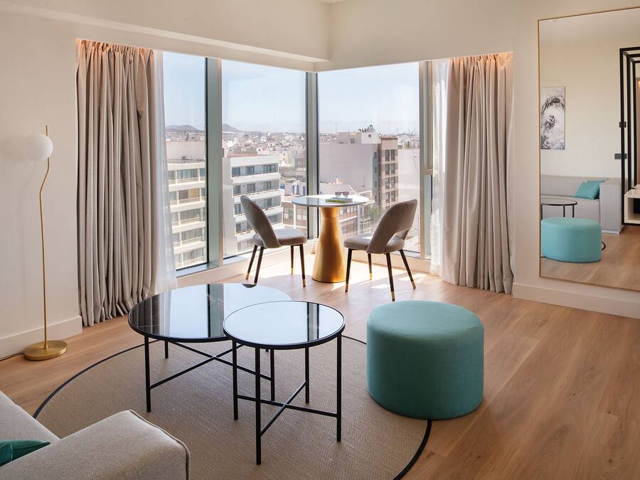 A modern hotel room with a large window, a round table, two chairs, a teal ottoman, and a sofa.