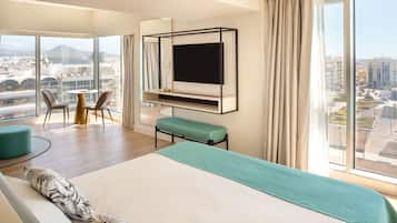 A modern hotel room with a large bed, a TV mounted on the wall, a seating area with chairs, and a view of the cityscape.