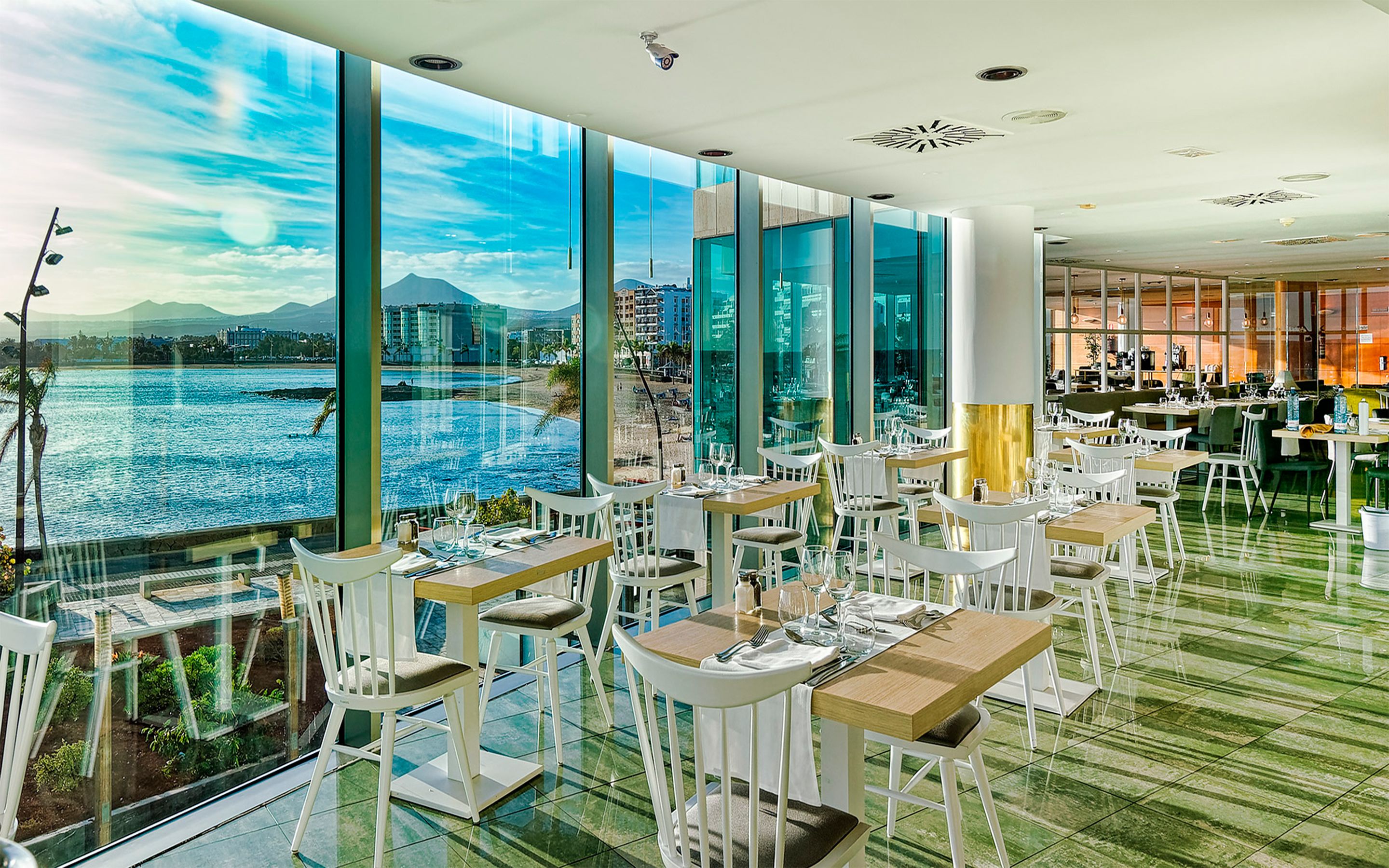 A restaurant with large windows offering a view of a body of water and a cityscape. Tables are arranged with white chairs, and the floor has a green tile pattern.