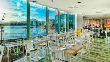 Un restaurante con grandes ventanales que ofrecen vistas al agua y al horizonte urbano. Las mesas están dispuestas con sillas blancas y el suelo presenta un diseño de baldosas verdes.