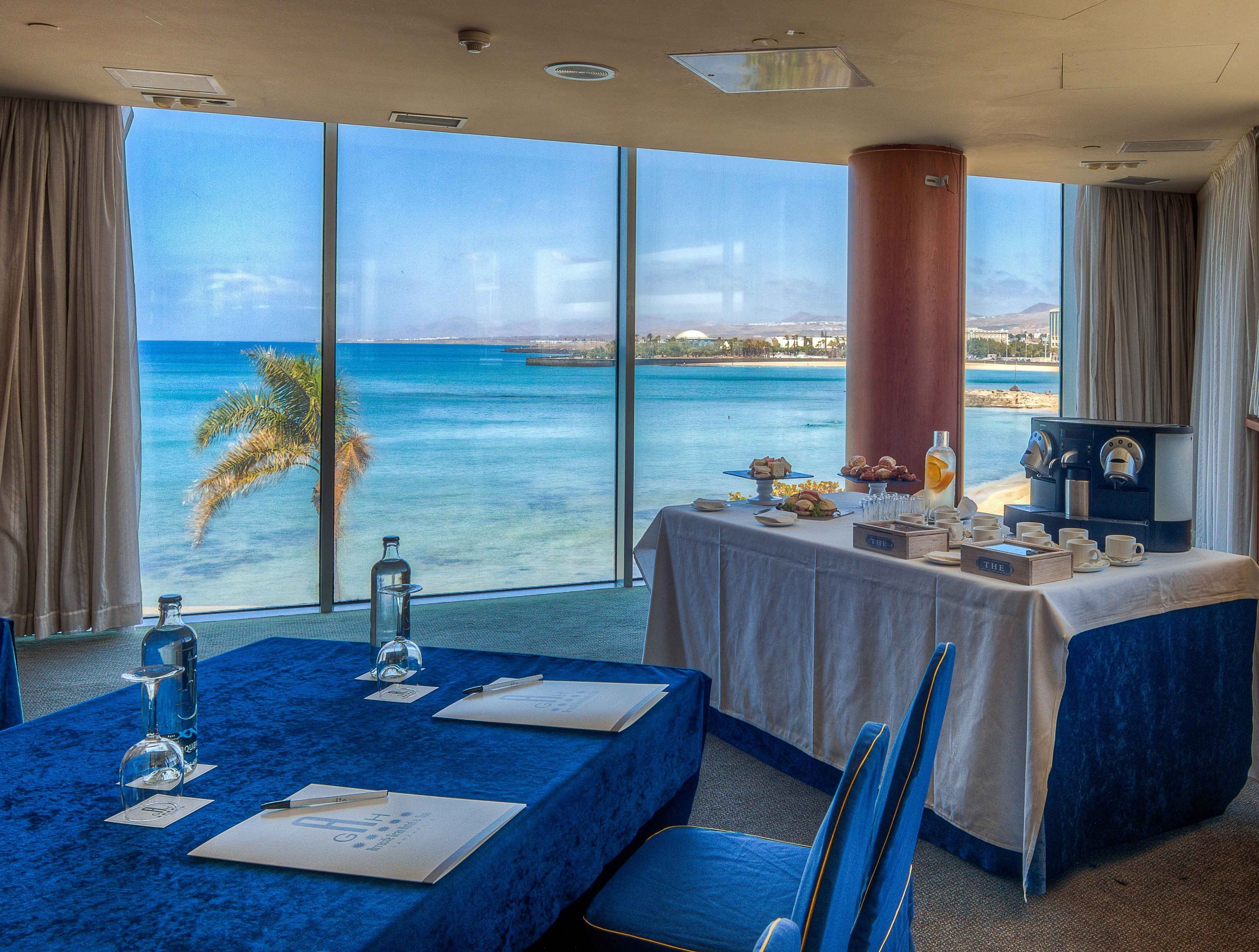 A room with a large window overlooking a beach, a table set with a coffee machine, and a view of the ocean.