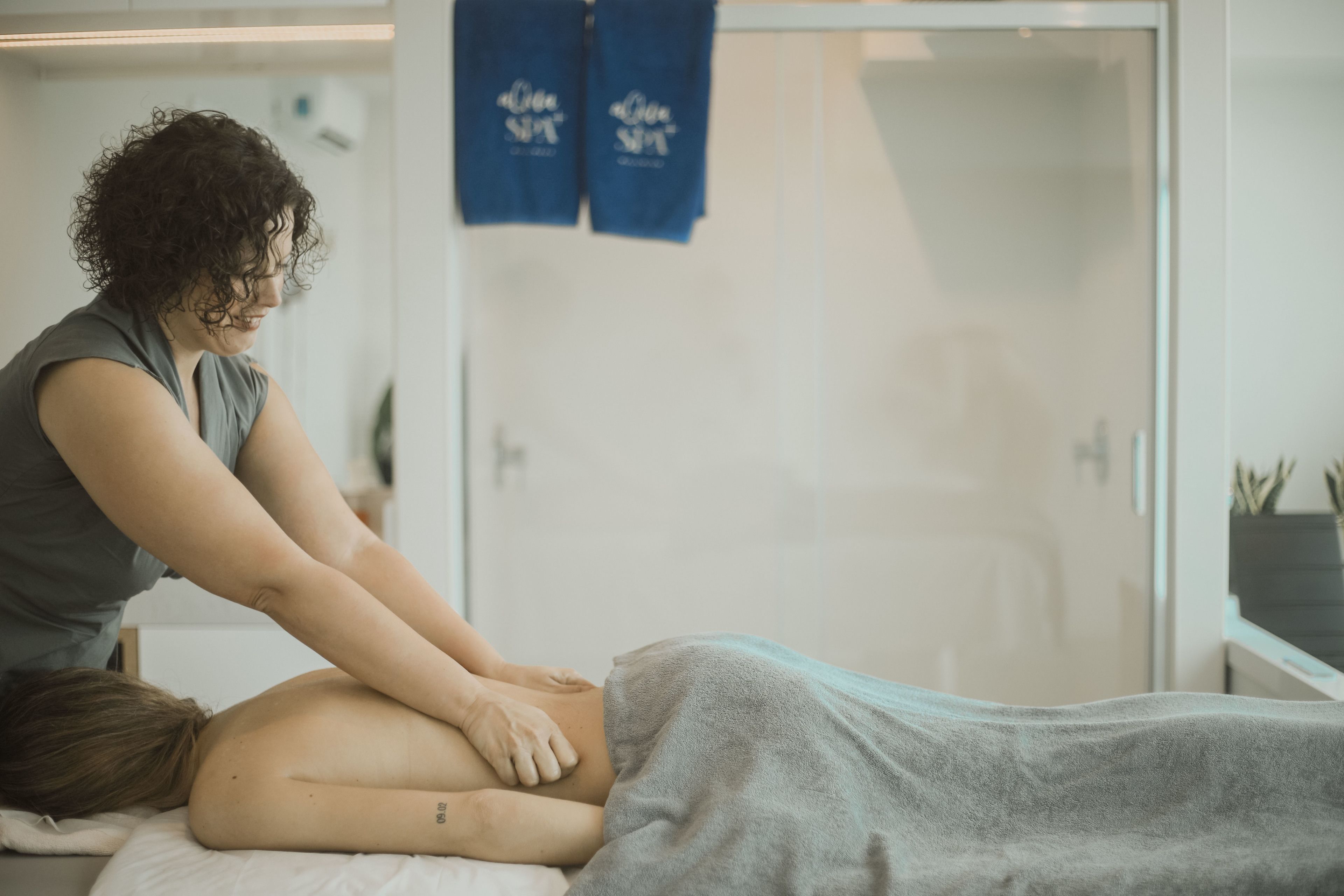 Sesión de terapia de masaje en un entorno clínico, con camilla, toallas y un separador de vidrio.