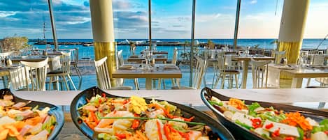 A restaurant with outdoor seating overlooking the sea, featuring multiple dining tables and chairs.