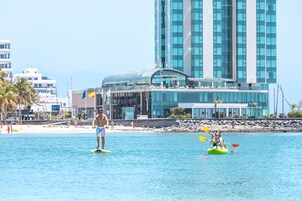 Un hotel moderno con fachada de vidrio, zona de piscina y playa al fondo.