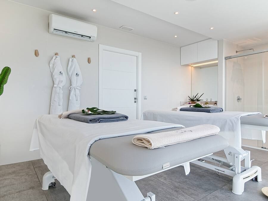Un spa moderno con dos camillas de tratamiento, un espejo grande, un cactus en maceta y una puerta blanca.