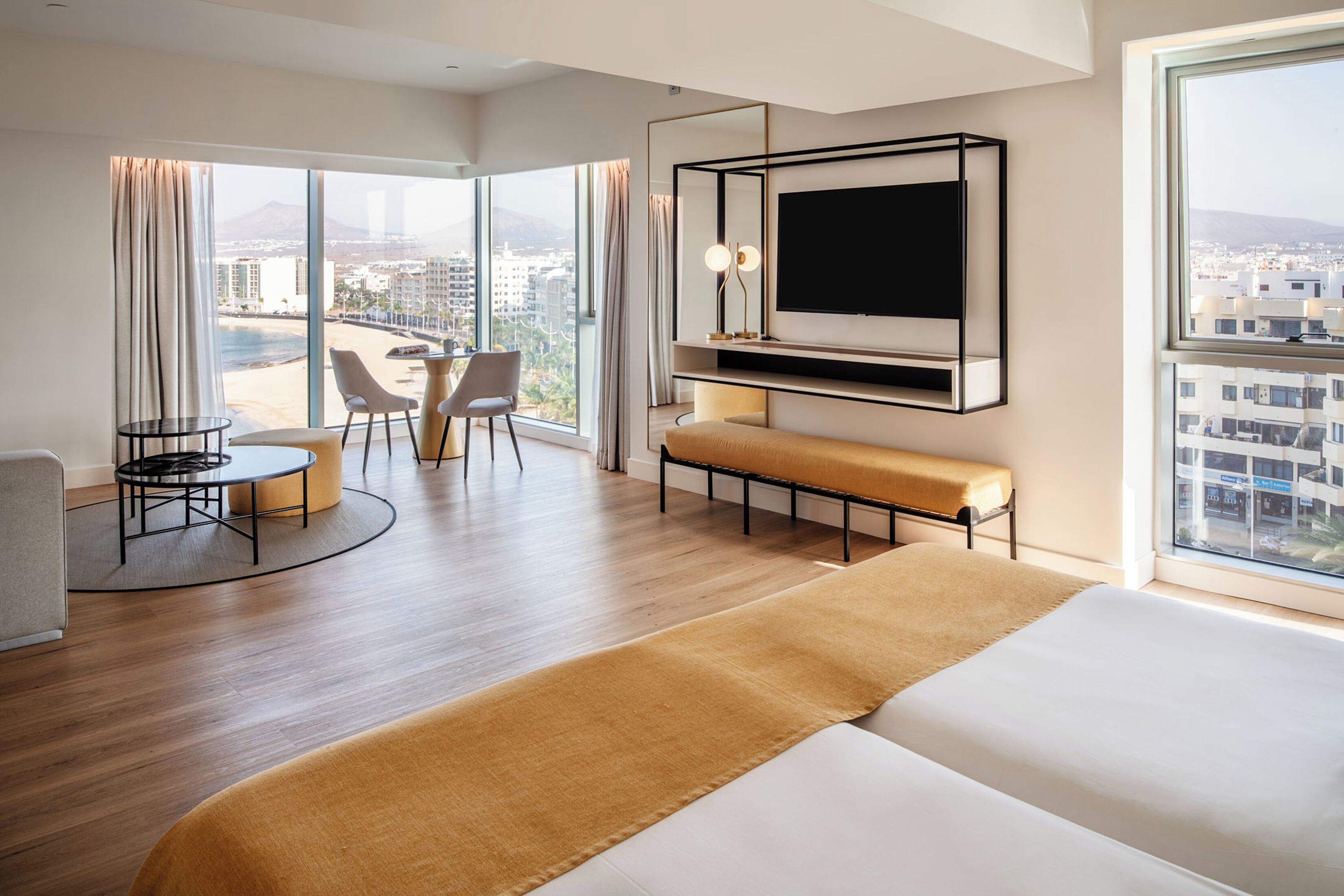 A modern hotel room with a large bed, a TV mounted on the wall, a seating area with a table and chairs, and a view of the city through large windows.