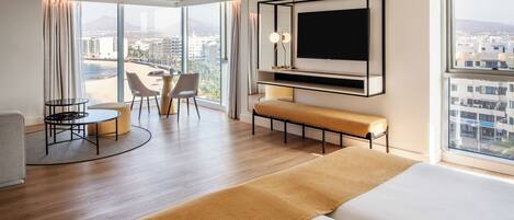 A modern hotel room with a large bed, a TV mounted on the wall, a seating area with a table and chairs, and a view of the city through large windows.