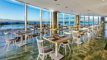 A restaurant with a view of the ocean, featuring tables and chairs arranged for dining.