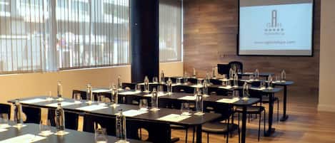A conference room with tables set for a meeting, a projector screen displaying a presentation, and large windows with blinds.