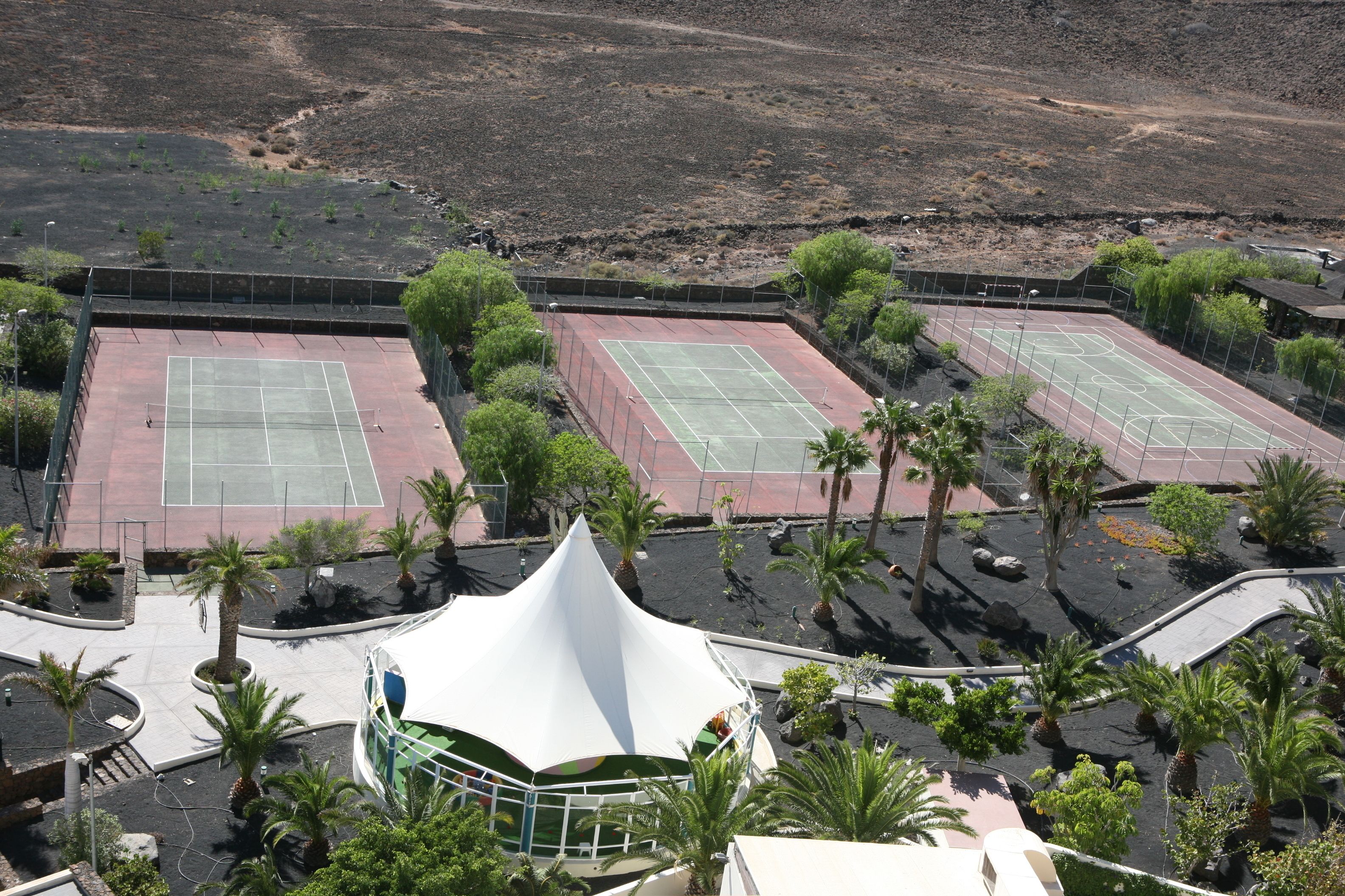 A tennis court complex with multiple courts, surrounded by palm trees and a white tent-like structure.